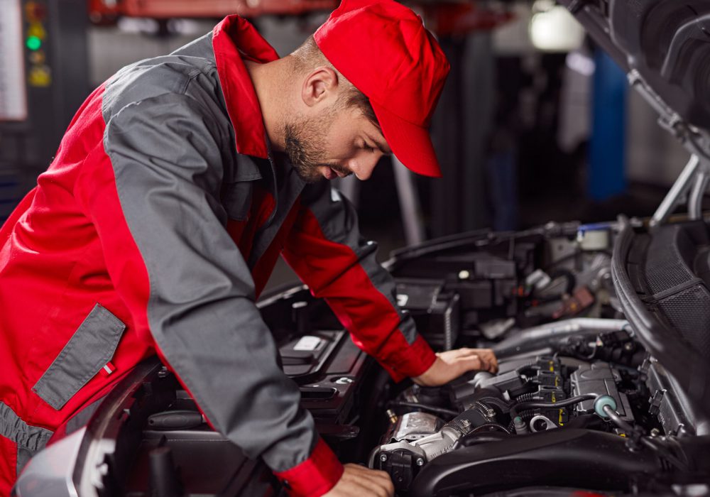 Side view of professional repairman in workwear standing near open hood of modern automobile and examining motor during diagnostic in workshop